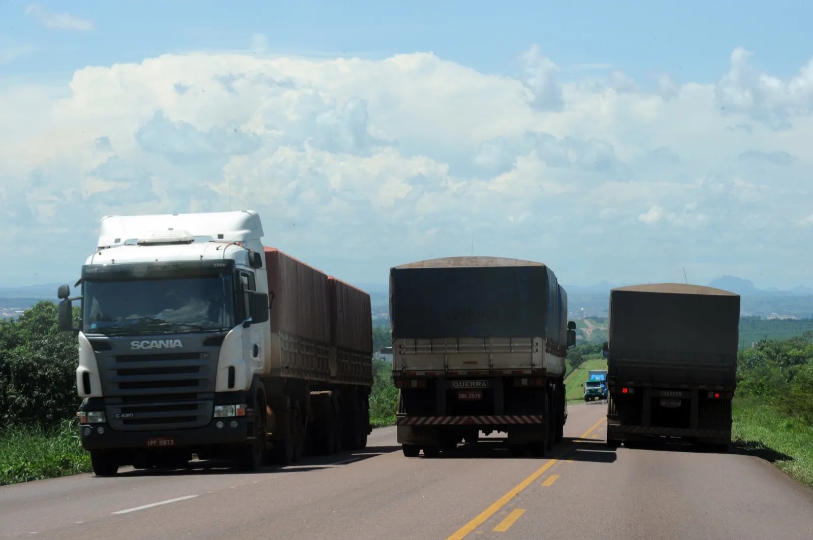 Três caminhões de carga coberta em uma rodovia de duas faixas, com paisagem rural e céu com nuvens ao fundo
