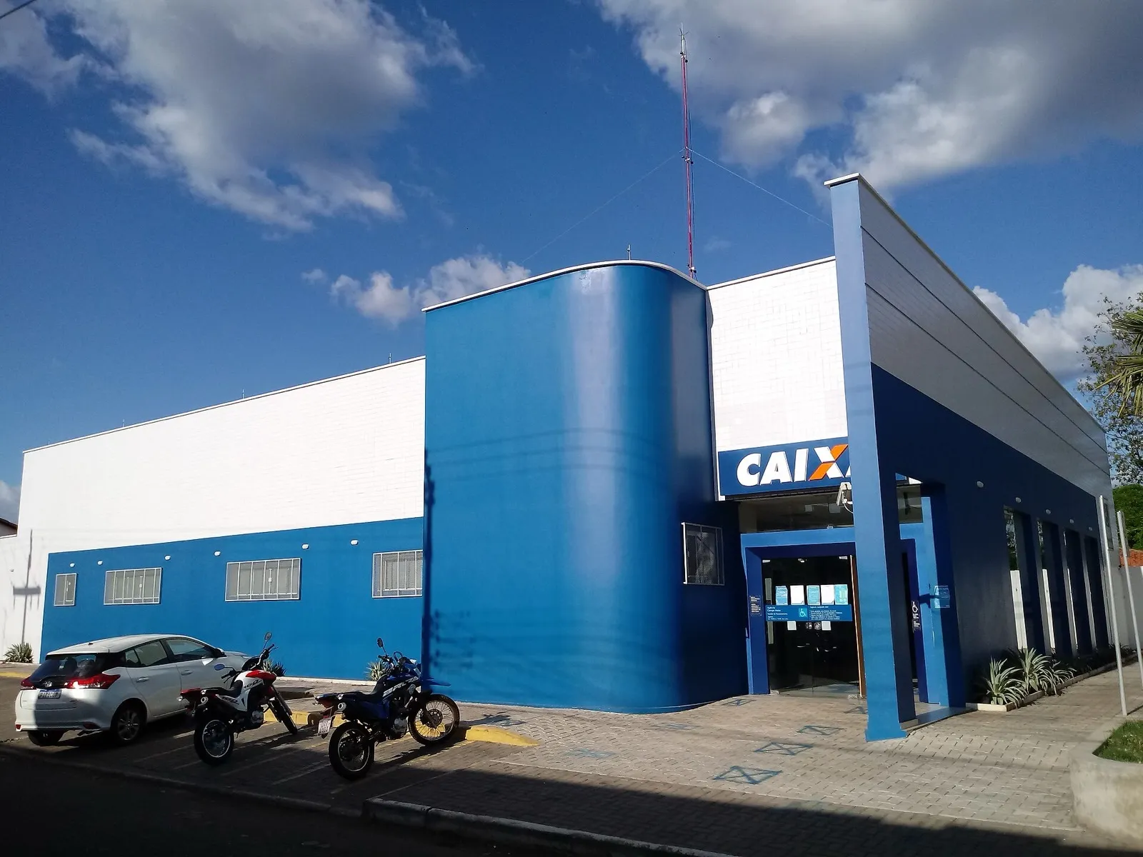 Fachada azul e branca de uma agência da Caixa Econômica Federal, com carro e motos estacionados em frente, sob céu azul com nuvens