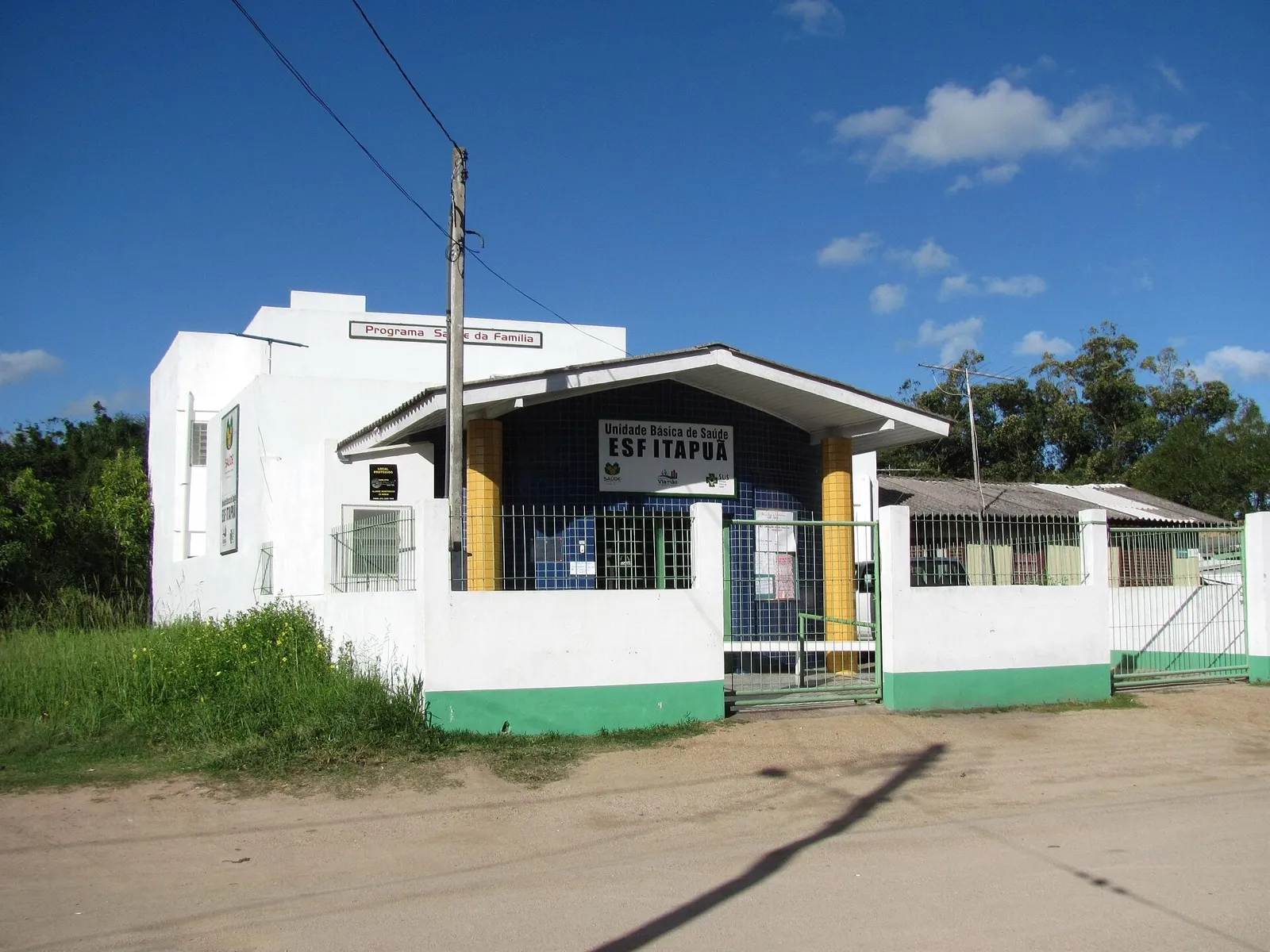Fachada de uma Unidade Básica de Saúde com placa da Estratégia Saúde da Família, muro branco e verde e portão gradeado, sob céu azul