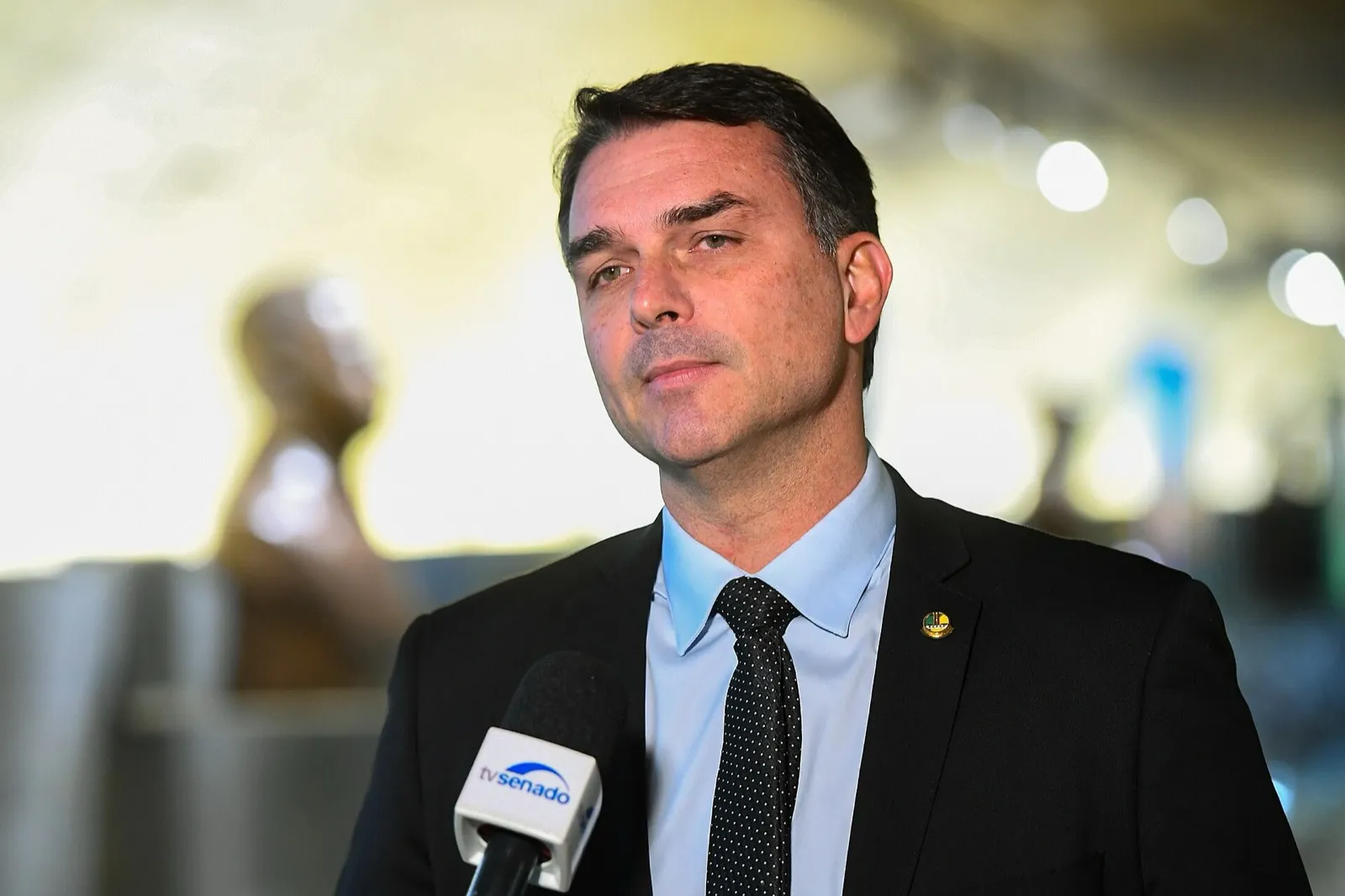 Senador Flávio Bolsonaro, de terno escuro e gravata, fala diante de um microfone da TV Senado