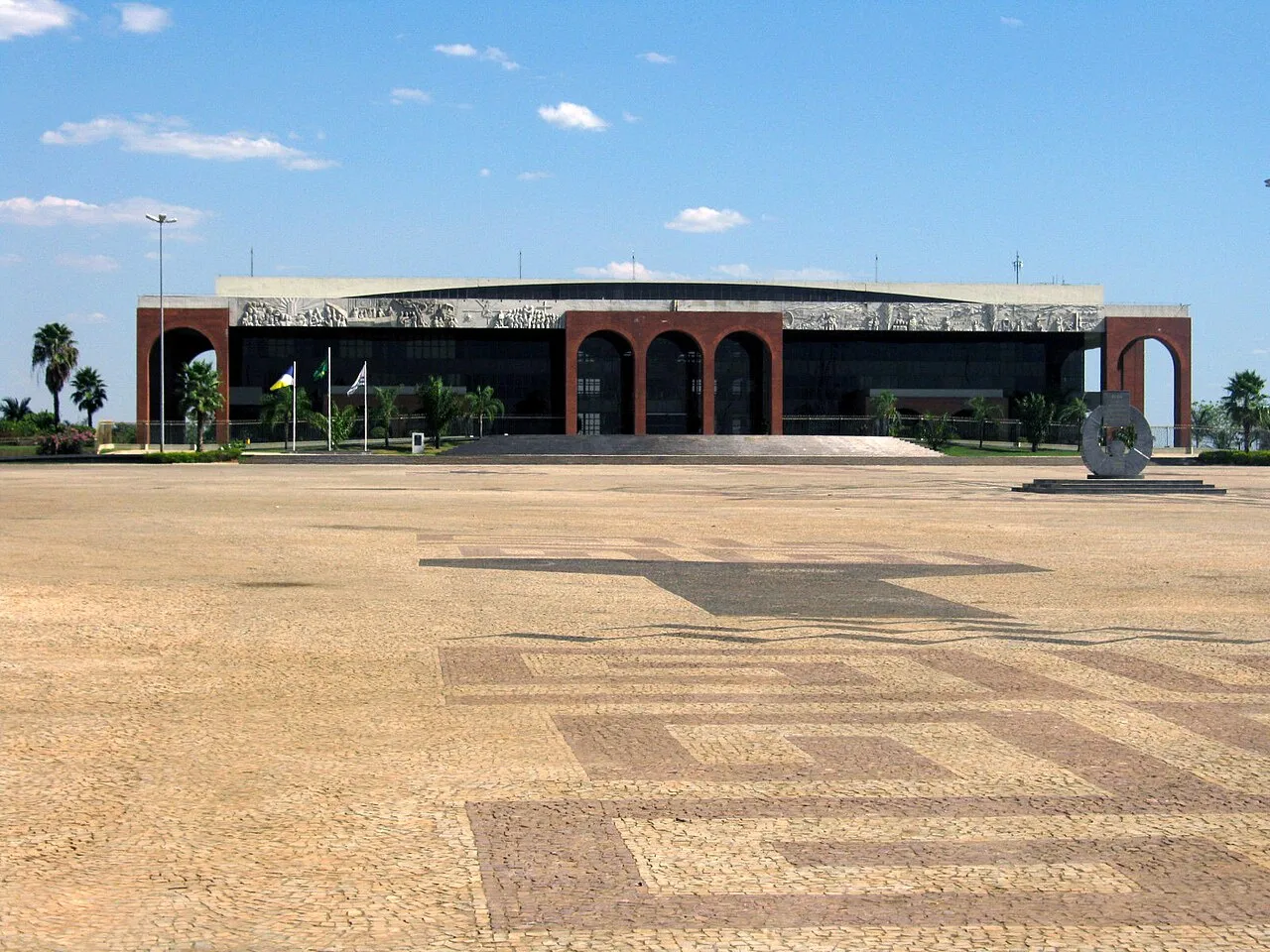Prédio horizontal de vidro escuro com arcos de tijolo vermelho e friso em relevo, bandeiras em mastros e uma grande praça calçada em primeiro plano