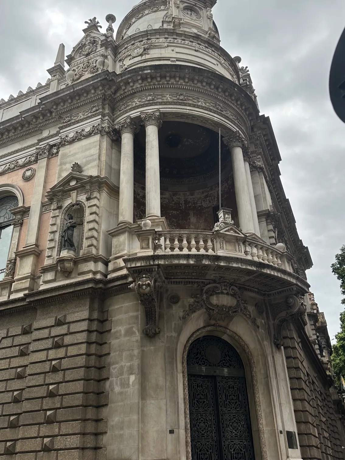 Fachada de prédio neoclássico com colunas, varanda e portas de bronze, sob céu nublado, em rua do Centro do Rio de Janeiro